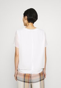 Haut blanc à manches courtes avec un panneau arrière transparent et un ourlet droit, assorti à un pantalon à carreaux aux motifs beiges et bruns.