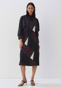 Schwarze kurze Jacke über einem midi Kleid mit buntem, geometrischem Muster in Burgunder- und Cremetönen, ergänzt durch Riemchensandalen.