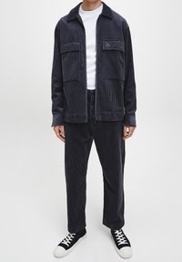 Marineblaue Cordjacke mit zwei Vordertaschen, kombiniert mit passenden Hosen und weißem T-Shirt mit Rundhalsausschnitt. Getragen mit schwarzen Sneakers.