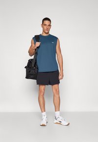 Teal sleeveless shirt, black shorts, white sneakers with gum soles, holding a black duffel bag. Minimal design, lightweight fabric.