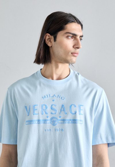 Helehall puuvillane T-särk, millel on tumedam sinine "VERSACE" logo, lühikesed varrukad, ümmargune kaelus ja allservas aastaarv "EST. 1978".