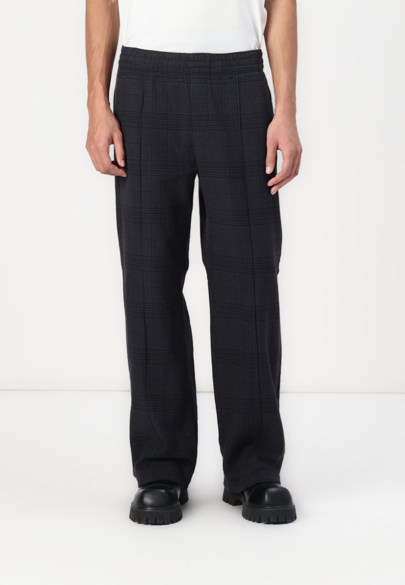 Homme portant un pantalon large à carreaux noirs avec une taille élastique et des chaussures noires épaisses, debout sur fond blanc.
