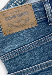 Traperice s svijetloplavim pranjem, s smešnim kožnim oznakom označenom "Next Denim Supply" i vidljivim detaljima šivanja.