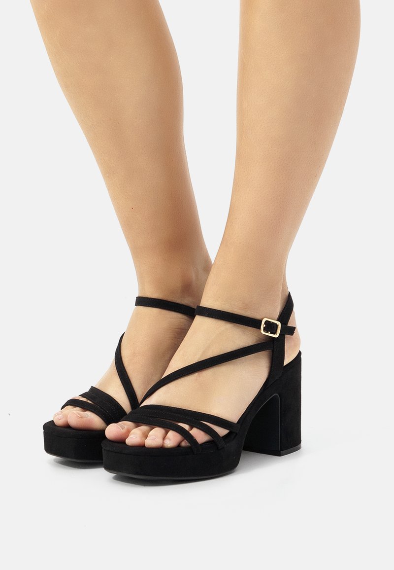 Sandalias de tacón de ante negro con múltiples tiras delgadas y un tacón cuadrado robusto. Detalle de hebilla en la correa del tobillo, diseño de punta abierta.