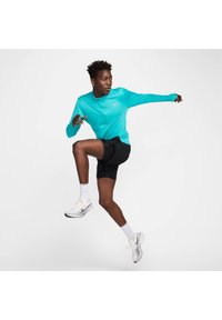 Langharige, turquoise sporttop met rits, gecombineerd met zwarte shorts en witte sneakers, met grijze accenten en veters.