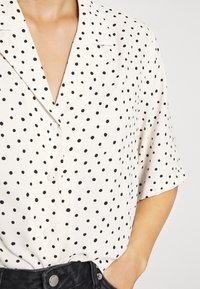 Camisa de botones de manga corta en tejido ligero color crema con un patrón de lunares negros; presenta un escote en V y un corte relajado.