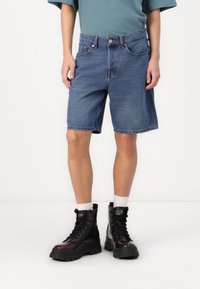 Shorts de mezclilla de un azul medio con un corte relajado, cinco bolsillos y costuras visibles. Combinados con botas negras gruesas y calcetines blancos hasta la mitad de la pierna.