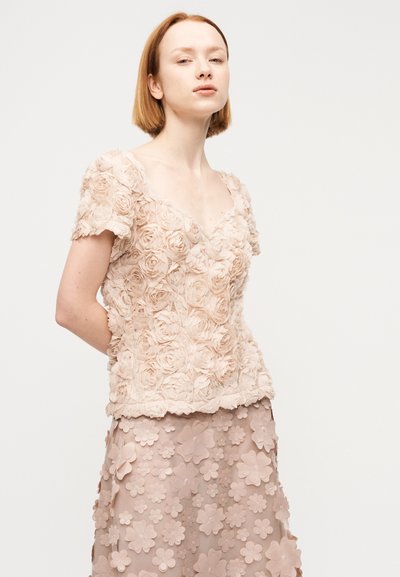 Νεαρή γυναίκα με κοντά κόκκινα μαλλιά φοράει μια υφασμάτινη μπεζ μπλούζα με τρισδιάστατα τριαντάφυλλα και μια ταιριαστή φλοράλ φούστα, σε απλό ανοιχτό φόντο.