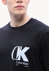 Musta puuvillapaita, jossa on pyöreä kaula-aukko. Paidassa on valkoinen "CK"-logo ja "Calvin Klein EST 1968" -teksti paksulla fontilla.