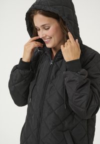 Veste noir matelassée avec capuche, dotée d'une fermeture éclair, de poignets côtelés et d'une surface texturée. Design simple et fonctionnel.