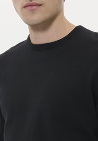 Svart stickad tröja med rund halsringning, med en slät textur och ribbad krage, visad från bröstet och uppåt mot en vit bakgrund.