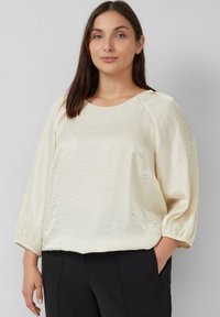 Blusa di un leggero avorio con una finitura testurizzata, scollatura rotonda e maniche a palloncino di tre quarti. Abbinata a pantaloni scuri.