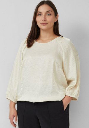 Helle Elfenbeintbluse mit strukturierter Oberfläche, rundem Ausschnitt und dreiviertellangen Ballonärmeln. Kombiniert mit dunklen Hosen.