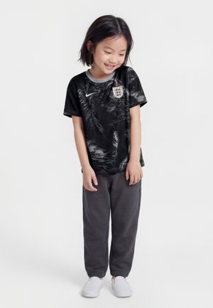 Jaunas vaikas šypsosi, dėvi juodą Anglijos futbolo marškinėlį, tamsiai pilkas kelnes ir baltus slip-on batelius, stovi priešais paprastą baltą foną.