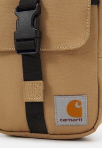 Saco Carhartt bege em tecido texturizado, com alça preta e fivela de plástico. Apresenta um rótulo cinza com um logótipo laranja e detalhes de costura.