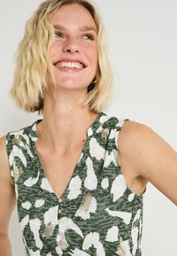 Top sin mangas verde con un patrón de leopardo, detalles en blanco y dorado, escote en V y tres botones en la parte delantera.