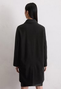 Cappotto in lana nera con una vestibilità rilassata, colletto a punta, maniche lunghe e silhouette snella. Texture liscia e dettagli minimi.
