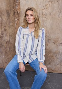 Jeune femme aux longs cheveux blonds assise sur une chaise, portant une chemise rayée crème et bleue et un jean bleu à jambes larges, devant un arrière-plan en bois.