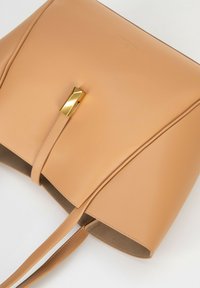 Borsa a mano in pelle beige con un design strutturato, dotata di una chiusura geometrica dorata e di spallacci sottili, presenta una texture liscia e cuciture minime.
