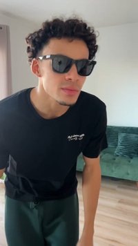 Zwart t-shirt met wit logo, zwarte zonnebril en donkergroene broek. Krullend haar en een neutrale gezichtsuitdrukking terwijl hij/zij voorover leunt.