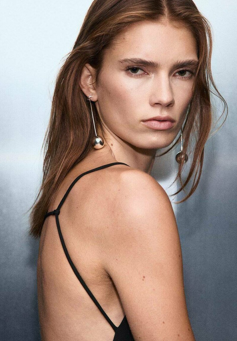 Svart klänning med spaghetti-remmar och öppen rygg, kombinerad med långa silvriga örhängen med stora sfäriska accenter. Slät textur och naturligt hår.