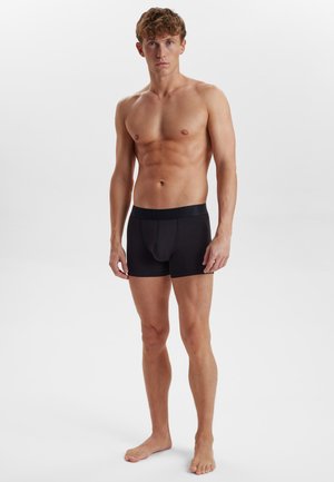 Schwarze Boxershorts mit strukturiertem Bund, figurbetontem Design und glatter Oberfläche. Das Modell steht barfuß vor einem weißen Hintergrund.