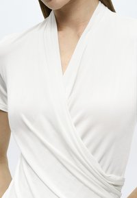 Λευκό κορμάκι τύπου wrap με κοντά μανίκια, κατασκευασμένο από μαλακό ύφασμα. Διαθέτει λαιμόκοψη σε σχήμα V και σχεδιασμό με drape, που αναδεικνύει μια απαλή υφή.