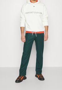 Vit hoodie med logotyp, röd innerfoder, gröna byxor och svarta sneakers med orangea snören. Avslappnad passform, mjukt tyg och dragsko i midjan.