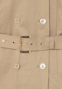 Béžový trench coat se zlatými akcenty, dvouřadý design a pásek. Obsahuje kulaté knoflíky a oka na pásku. Hladká textura.
