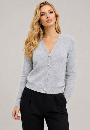 Femme blonde portant un cardigan gris clair avec des boutons en perle et un pantalon noir, posant avec une main partiellement dans la poche sur un fond uni.