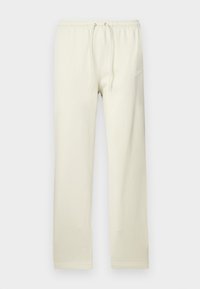 Calças de treino bege claro com cintura ajustável por cordão, feitas de material macio. Apresentam um discreto logótipo Nike branco na coxa esquerda.