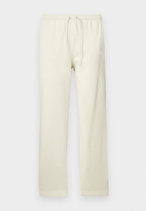 Calças de treino bege claro com cintura ajustável por cordão, feitas de material macio. Apresentam um discreto logótipo Nike branco na coxa esquerda.