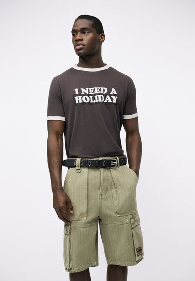 Άνδρας που φοράει σκουρόχρωμο μπλουζάκι με το κείμενο «ΧΡΕΙΑΖΟΜΑΙ ΔΙΑΚΟΠΕΣ», συνδυασμένο με ανοιχτόχρωμο ελαιοπράσινο cargo σορτς και μαύρη ζώνη, στέκεται μπροστά σε λευκό φόντο.
