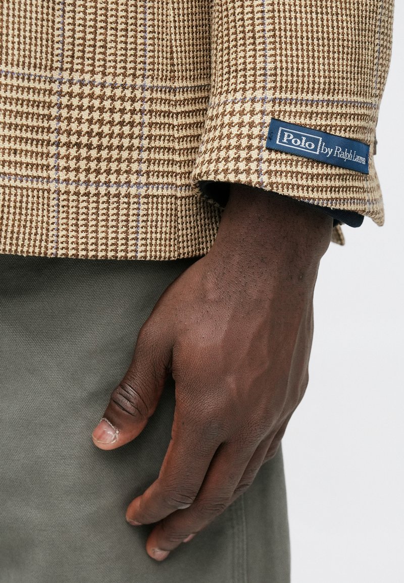 Brown houndstooth blazer with a textured finish and blue "Polo by Ralph Lauren" label. Visible hand is resting on an olive-green fabric.