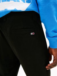 Zwarte broek met een gladde textuur, voorzien van een achterzak en een klein geborduurd logo in rood, wit en blauw. Blauwe sweater zichtbaar erboven.
