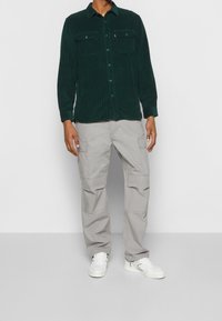 Zielona sztruksowa koszula z długim rękawem i dwiema kieszeniami na piersi, w połączeniu z jasnoszarymi spodniami cargo z wieloma kieszeniami po bokach i białymi trampkami.