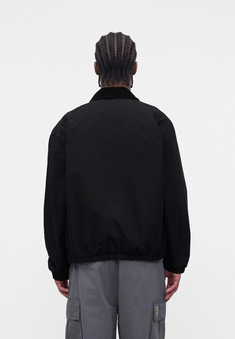Person mit geflochtenem Haar, die eine schwarze Jacke und graue Cargohosen trägt, steht mit dem Rücken zu einem einfarbigen hellen Hintergrund.
