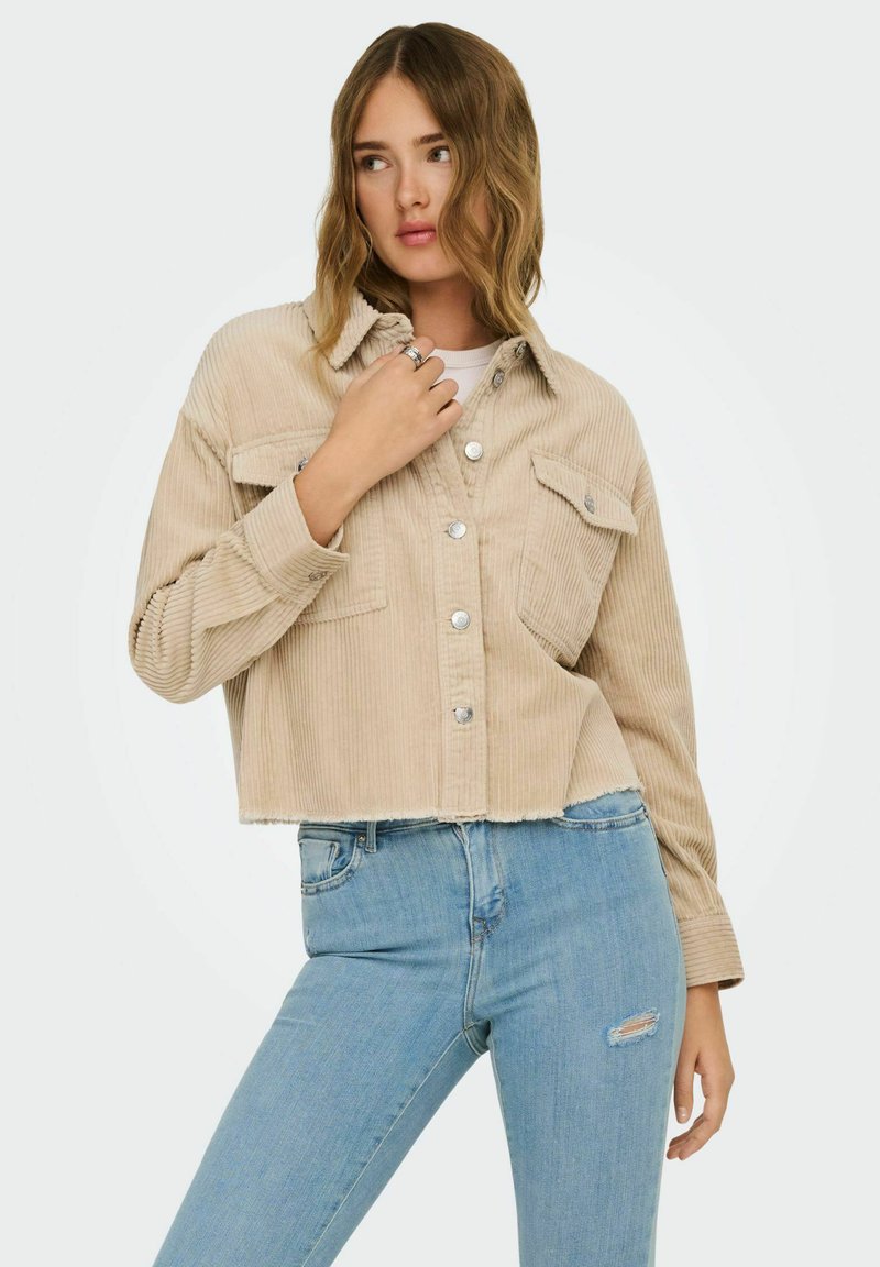 Nő, aki bézs színű kordbársony rövidített dzsekit visel ezüstszínű gombokkal és világoskék, szakadt szűk farmernadrággal, miközben a gallér közelében tartja a kezét.