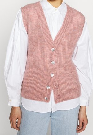 Różowa dzianinowa kamizelka bez rękawów z prążkowanym wzorem i pięcioma ozdobnymi guzikami, noszona na białej koszuli zapinanej na guziki z bufkami na długich rękawach oraz jasnoniebieskich dżinsach.