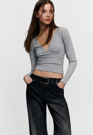 Giovane donna con lunghi capelli scuri indossa una maglia grigia a portafoglio con maniche lunghe e pantaloni neri a vita alta con cintura, mani nelle tasche.