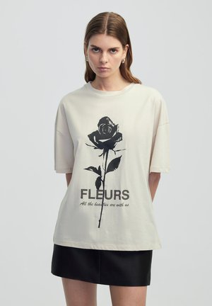T-shirt oversize beige clair en coton. Présente un graphique de rose noire et le texte "FLEURS" avec "Toutes les beautés sont avec nous" en dessous.