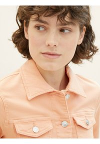 Veste en denim pêche avec un col pointu, des fermetures à boutons sur le devant et deux poches poitrine avec des accents métalliques. Texture douce et design ajusté.