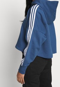 Blå kortärmad hoodie gjord av mjukt material, med en stor huva och vita randdetaljer på ärmarna. Matchad med svarta byxor.