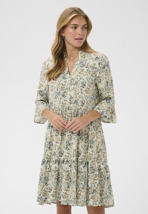 Kobieta w długiej, sięgającej kolan sukience w kwiaty w niebiesko-brązowe wzory, stojąca z złączonymi rękami i patrząca w bok.