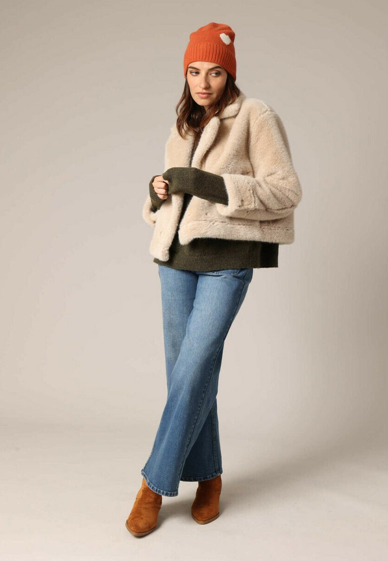 Crème nepleren jas met olijfgroene gebreide manchetten, gecombineerd met een donkergroen sweater, blauwe flare jeans en een oranje muts. Bruine suède enkellaarzen.