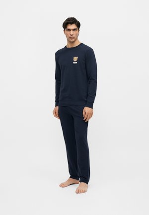 Férfi áll mezítláb, sötétkék hosszú ujjú felsőt és nadrágot visel, melyen kis medve grafika van a mellkasán.