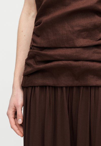 Καφέ τοπ με υφή στρογγυλεμένου υφάσματος, σχεδιασμό με στρώσεις και ρομαντική σιλουέτα, συνδυασμένο με αέρινη καφέ φούστα με πτυχώσεις.