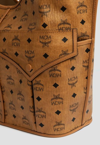 Skórzana torba typu tote w kolorze tan, ozdobiona wytłoczonymi czarnymi logo MCM oraz kształtami w diamenty. Zawiera przednią kieszeń z zapięciem na zatrzask oraz teksturowane wykończenie.