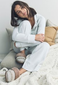 Set da casa grigio chiaro con tessuto morbido, composto da una maglia a maniche lunghe e pantaloni larghi. Pantofole marroni e grigie senza lacci con bordo in pelliccia.