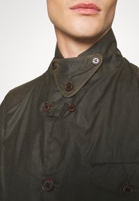 Dark green jacket with a high collar, featuring brown buttons and a side pocket. The fabric appears textured and slightly glossy.